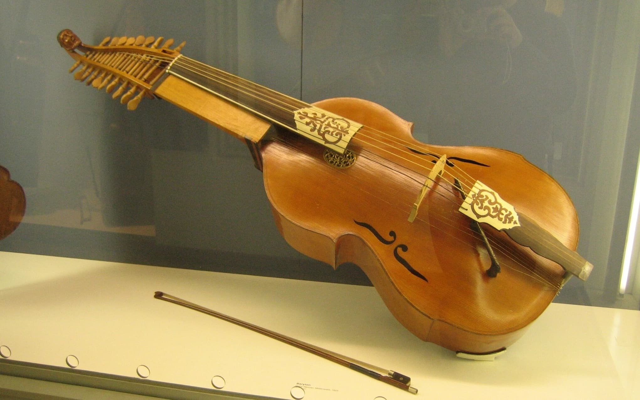 Baryton in display case, showing six bowable strings and twelve sympathetic strings beneath them. Also shown is a bow.