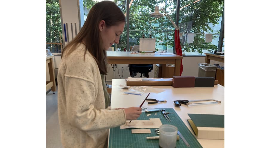 Abigail Mead standing at a desk working on a copy of a 19th century stationary binding using conservation tools.