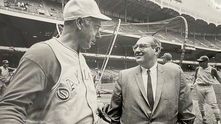 Two men facing each other, the one of the right in a business suit smiling at the man on the left who is wearing a baseball uniform.
